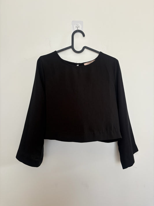 The Circle black cropped blouse with long bell sleeves and a round neckline hanging on a hanger against a white wall.