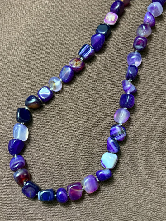 The Circle purple agate beaded necklace featuring smooth tumbled stones with natural banding on a brown fabric background