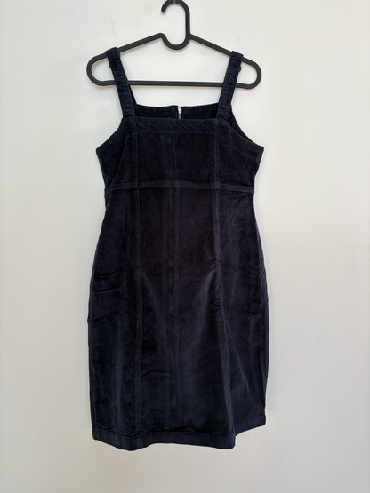 The Circle black corduroy sleeveless pinafore dress with a square neckline and front zipper, hanging on a hanger.