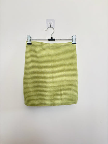 The Circle lime green mini skirt in a textured waffle-knit fabric hanging on a metal hanger against a white wall.