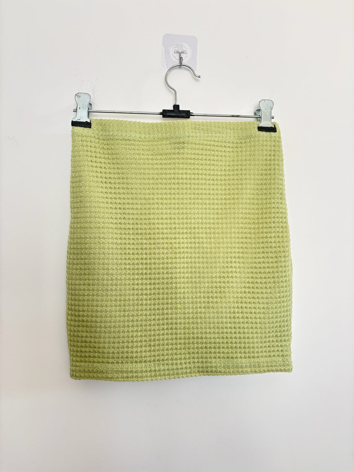 The Circle lime green mini skirt in a textured waffle-knit fabric hanging on a metal clip hanger against a white wall.