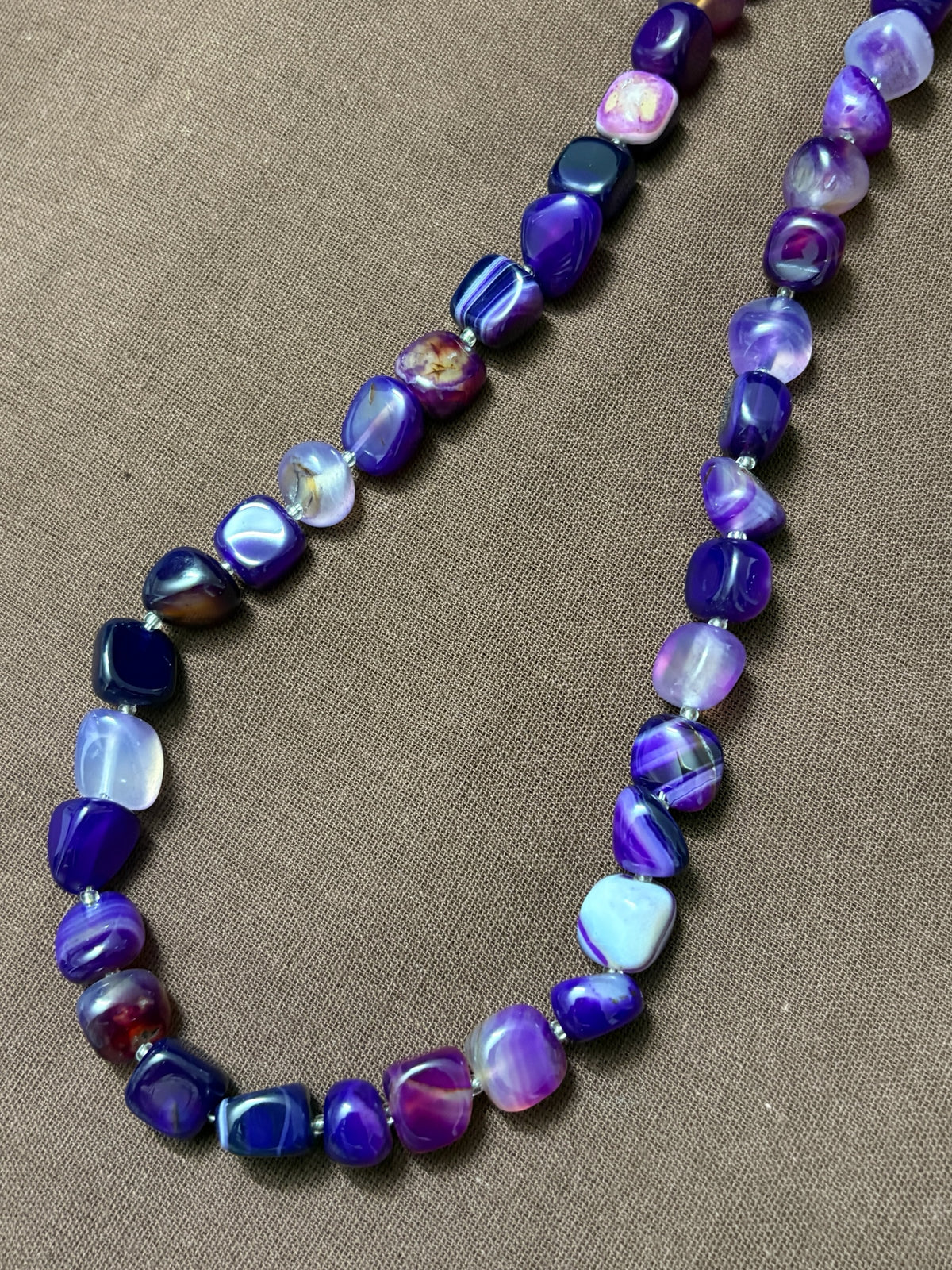 The Circle purple agate beaded necklace featuring smooth tumbled stones with natural banding on a brown fabric background