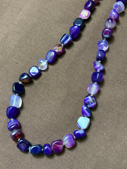 The Circle purple agate beaded necklace featuring smooth tumbled stones with natural banding on a brown fabric background